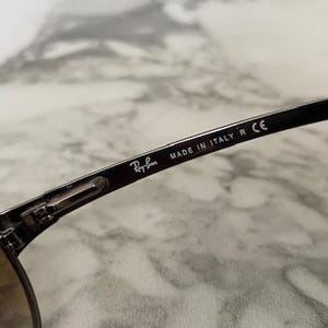 May include: Close-up of Ray-Ban sunglasses. The black frame features the Ray-Ban logo and the text "MADE IN ITALY R CE". The lenses are a gradient brown color. The sunglasses are on a marble surface.