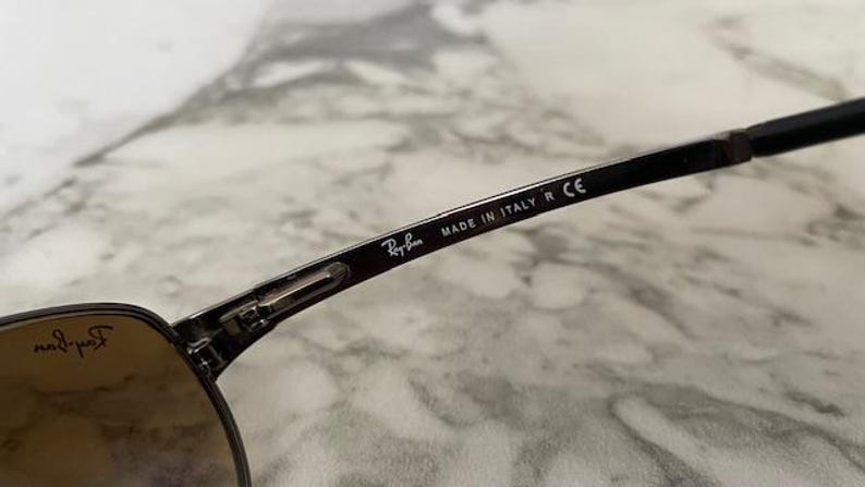 May include: Close-up of Ray-Ban sunglasses. The black frame features the Ray-Ban logo and the text "MADE IN ITALY R CE". The lenses are a gradient brown color. The sunglasses are on a marble surface.