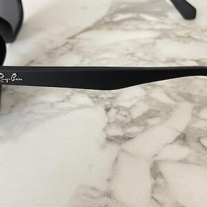 Pu&ograve; includere: Occhiali da sole neri con lenti scure. Il logo Ray-Ban &egrave; visibile sull'asta. Gli occhiali da sole sono appoggiati su uno sfondo di marmo.