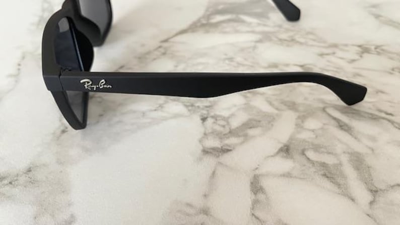 Pu&ograve; includere: Occhiali da sole neri con lenti scure. Il logo Ray-Ban &egrave; visibile sull'asta. Gli occhiali da sole sono appoggiati su uno sfondo di marmo.