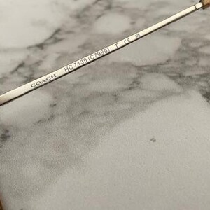 May include: Close-up of Coach sunglasses. The silver metal arm features the brand name and model number HC 7135 (C7999). The lenses are a brown tint. The sunglasses are set against a marble background.