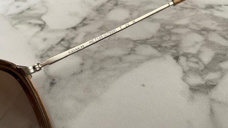 May include: Close-up of Coach sunglasses. The silver metal arm features the brand name and model number HC 7135 (C7999). The lenses are a brown tint. The sunglasses are set against a marble background.