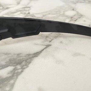 Puede incluir: Primer plano de gafas de sol negras con lentes azules. Las gafas de sol tienen un dise&ntilde;o elegante y moderno con una patilla curvada. La montura es de color negro mate y las lentes son de color azul vibrante.