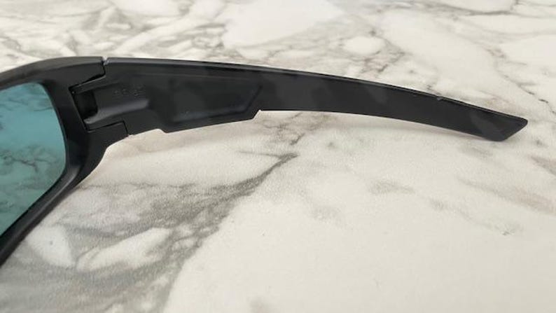Puede incluir: Primer plano de gafas de sol negras con lentes azules. Las gafas de sol tienen un dise&ntilde;o elegante y moderno con una patilla curvada. La montura es de color negro mate y las lentes son de color azul vibrante.
