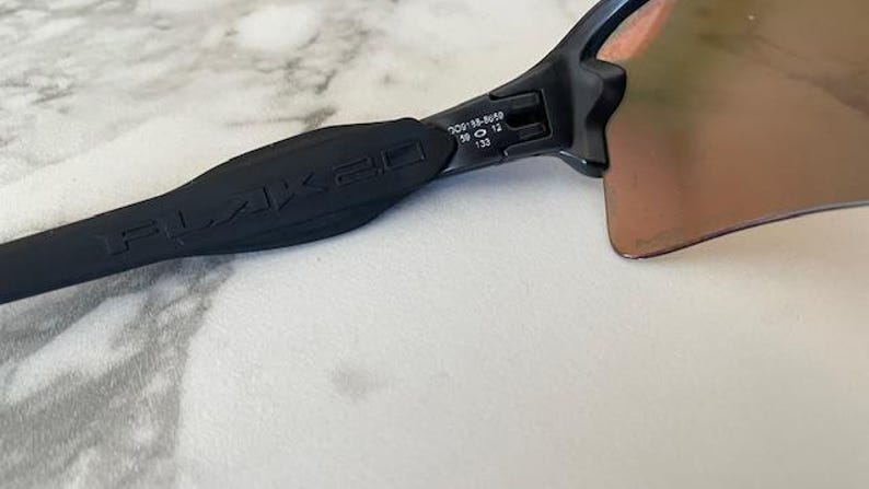Peut inclure: Gros plan sur des lunettes de soleil noires avec le logo FLAK 2.0 sur la branche. Les verres sont de couleur marron d&eacute;grad&eacute;. La monture porte les chiffres 58 12 et 133 sur la branche.