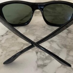 Puede incluir: Gafas de sol negras con lentes verde oscuro. Las gafas tienen una montura rectangular gruesa y patillas que se cruzan en el centro. Las lentes son de color verde oscuro y la montura es negra. Sobre un fondo de m&aacute;rmol.