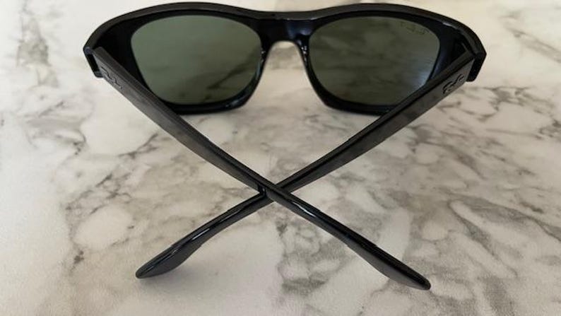 Puede incluir: Gafas de sol negras con lentes verde oscuro. Las gafas tienen una montura rectangular gruesa y patillas que se cruzan en el centro. Las lentes son de color verde oscuro y la montura es negra. Sobre un fondo de m&aacute;rmol.