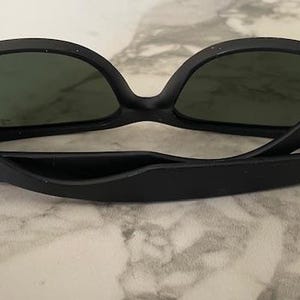 May include: Black sunglasses with dark green lenses. The sunglasses have a matte finish and the brand name is visible on the temples. The glasses are resting on a light-colored, marble-like surface.