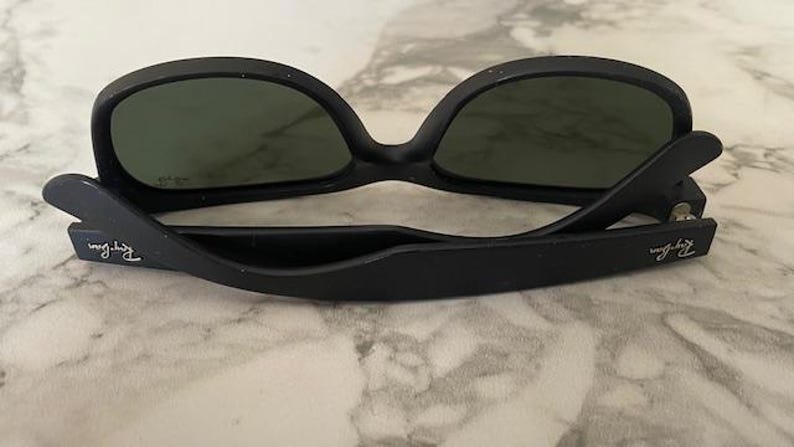 May include: Black sunglasses with dark green lenses. The sunglasses have a matte finish and the brand name is visible on the temples. The glasses are resting on a light-colored, marble-like surface.