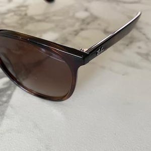 May include: Brown tortoiseshell sunglasses with dark lenses. The sunglasses have a classic design with a rectangular frame and a subtle logo on the temple. The lenses are a gradient brown color.