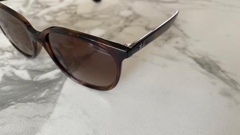 May include: Brown tortoiseshell sunglasses with dark lenses. The sunglasses have a classic design with a rectangular frame and a subtle logo on the temple. The lenses are a gradient brown color.