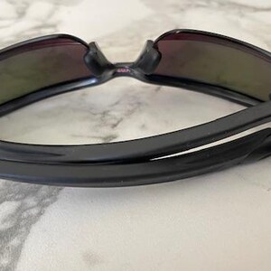 Puede incluir: Gafas de sol negras con montura curvada y lentes tintadas. Las gafas de sol est&aacute;n plegadas, mostrando las bisagras y el interior de las patillas. Las lentes tienen un tinte degradado, que pasa de un color m&aacute;s oscuro en la parte superior a un tono m&aacute;s claro.