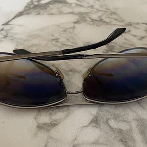 May include: Sunglasses with a silver metal frame and gradient lenses. The lenses transition from brown to blue, and the temples are black. The sunglasses are resting on a marble surface.