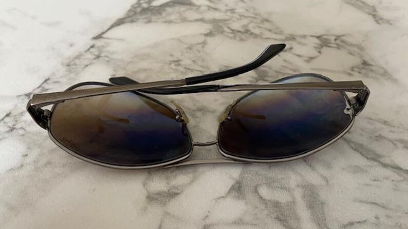 May include: Sunglasses with a silver metal frame and gradient lenses. The lenses transition from brown to blue, and the temples are black. The sunglasses are resting on a marble surface.