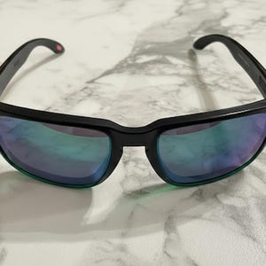 Puede incluir: Gafas de sol negras con montura rectangular y lentes iridiscentes. Las gafas de sol tienen un diseño elegante con un sutil acento verde en la parte inferior de las lentes. Las patillas tienen un pequeño logotipo.