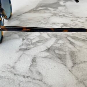 May include: Tortoiseshell-framed sunglasses with blue-tinted lenses. The sunglasses have a rounded shape and the Ray-Ban logo is visible on the temple. The frames are brown and black.