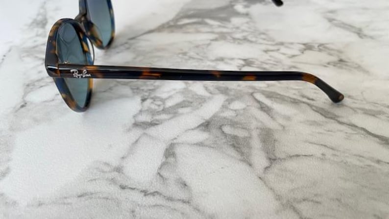 May include: Tortoiseshell-framed sunglasses with blue-tinted lenses. The sunglasses have a rounded shape and the Ray-Ban logo is visible on the temple. The frames are brown and black.
