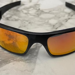 Puede incluir: Gafas de sol negras con lentes de color naranja. Las gafas tienen forma rectangular y una montura negra gruesa. Las lentes reflejan el entorno. Las gafas est&aacute;n sobre una superficie blanca y gris.