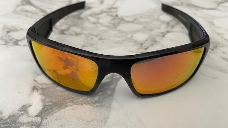 Puede incluir: Gafas de sol negras con lentes de color naranja. Las gafas tienen forma rectangular y una montura negra gruesa. Las lentes reflejan el entorno. Las gafas est&aacute;n sobre una superficie blanca y gris.