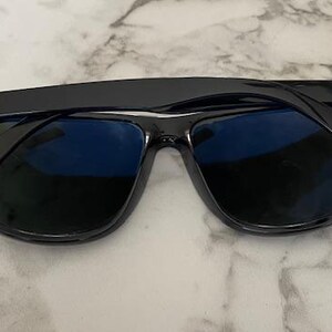 Puede incluir: Gafas de sol negras con lentes azules, con el logotipo de Ray-Ban en las patillas. Las gafas de sol est&aacute;n sobre un fondo de m&aacute;rmol. Las monturas son brillantes y reflejan la luz.