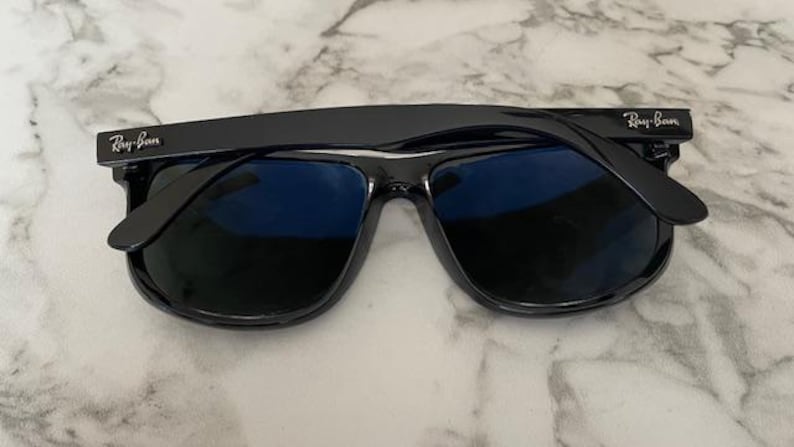 Puede incluir: Gafas de sol negras con lentes azules, con el logotipo de Ray-Ban en las patillas. Las gafas de sol est&aacute;n sobre un fondo de m&aacute;rmol. Las monturas son brillantes y reflejan la luz.