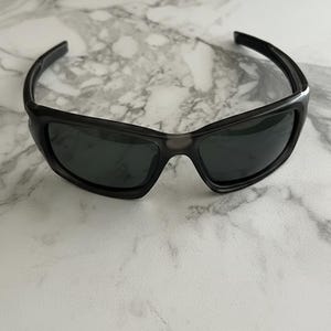 May include: Black sunglasses with a wraparound design. The sunglasses have dark lenses and a matte black frame. The image shows the sunglasses on a white and grey marble surface.