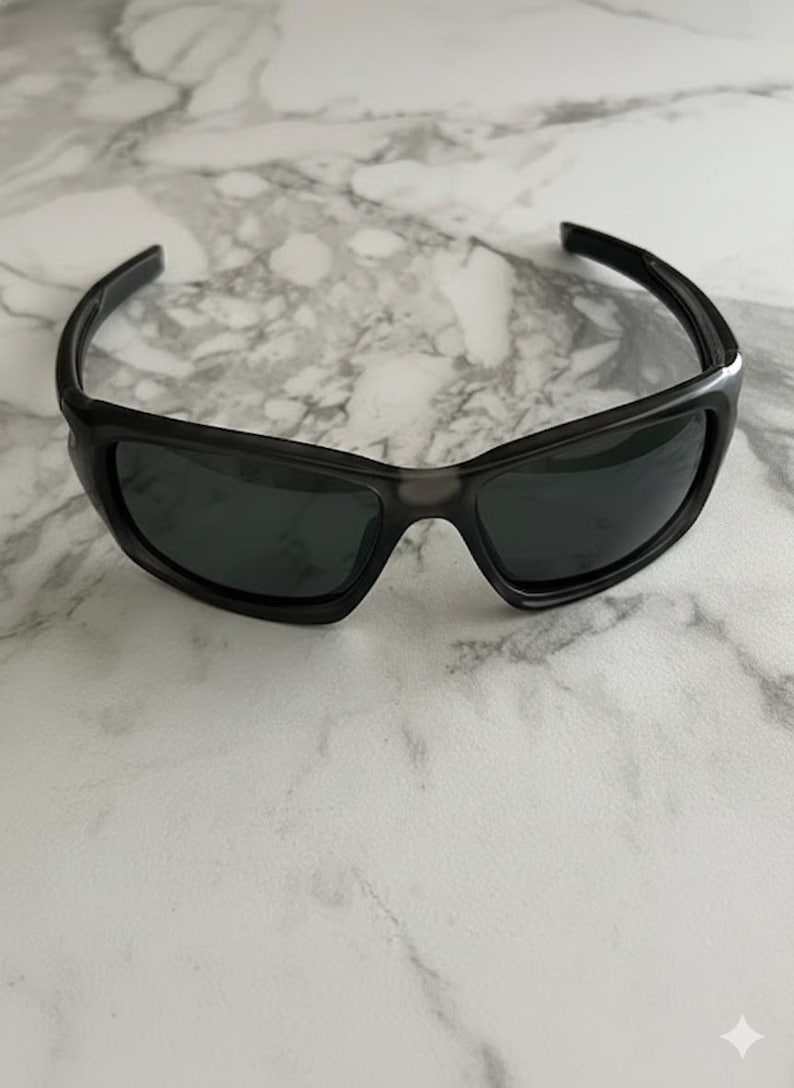 May include: Black sunglasses with a wraparound design. The sunglasses have dark lenses and a matte black frame. The image shows the sunglasses on a white and grey marble surface.