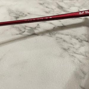 May include: Close-up of a pair of burgundy sunglasses. The frame has a rectangular shape with a dark lens. The arm of the glasses has the text "Hill Tribe" and other numbers.