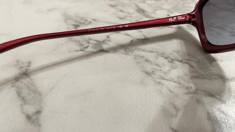 May include: Close-up of a pair of burgundy sunglasses. The frame has a rectangular shape with a dark lens. The arm of the glasses has the text "Hill Tribe" and other numbers.