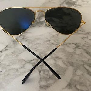 May include: Gold-framed aviator sunglasses with dark lenses. The sunglasses have a double bridge and black earpieces. The lenses reflect a blue hue. The sunglasses are set against a marble background.