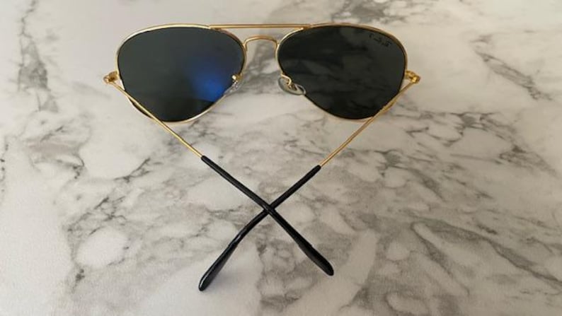 May include: Gold-framed aviator sunglasses with dark lenses. The sunglasses have a double bridge and black earpieces. The lenses reflect a blue hue. The sunglasses are set against a marble background.