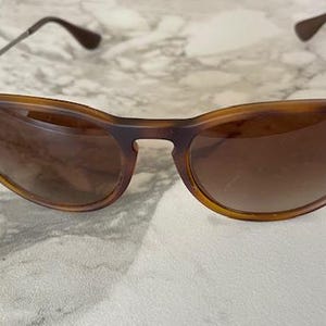 May include: Brown tortoiseshell sunglasses with a cat-eye shape. The sunglasses have brown lenses and silver-toned arms. The Ray-Ban logo is visible on the left lens. The sunglasses are on a marble surface.