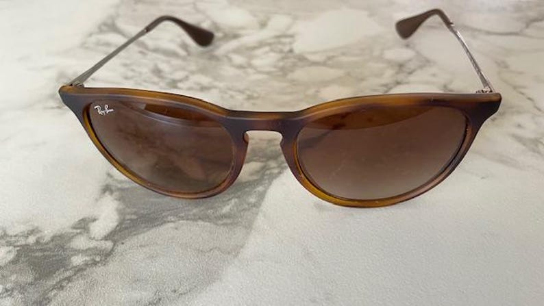 May include: Brown tortoiseshell sunglasses with a cat-eye shape. The sunglasses have brown lenses and silver-toned arms. The Ray-Ban logo is visible on the left lens. The sunglasses are on a marble surface.