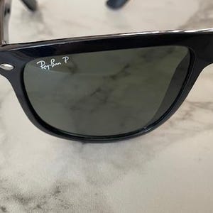 Puede incluir: Gafas de sol negras con lentes verdes. El logotipo de Ray-Ban es visible en la esquina superior izquierda de la montura. Las gafas de sol est&aacute;n sobre una superficie de m&aacute;rmol blanco y gris.