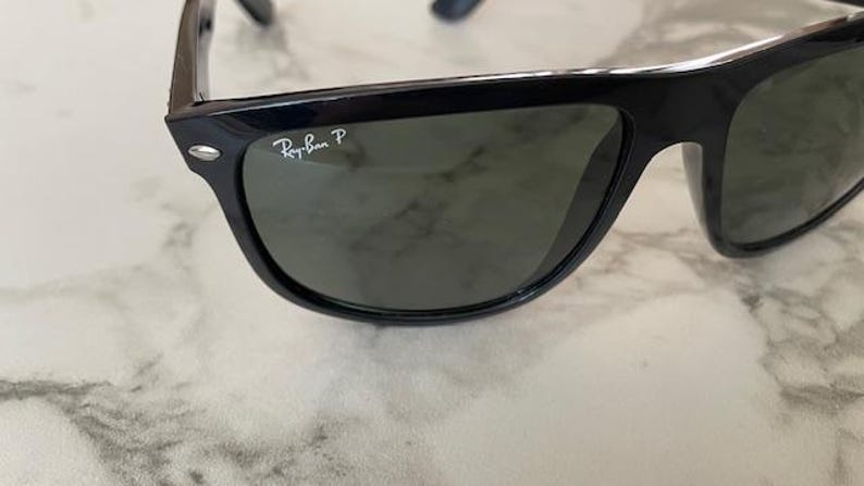 Puede incluir: Gafas de sol negras con lentes verdes. El logotipo de Ray-Ban es visible en la esquina superior izquierda de la montura. Las gafas de sol est&aacute;n sobre una superficie de m&aacute;rmol blanco y gris.
