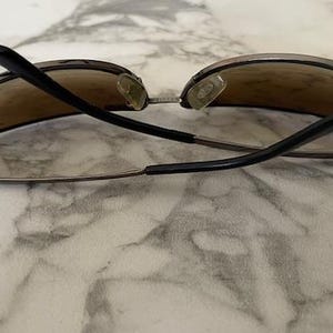 May include: Sunglasses with brown tinted lenses and black arms. The sunglasses have a silver frame and nose pads. The arms are curved and extend from the lenses. The sunglasses are resting on a marble surface.