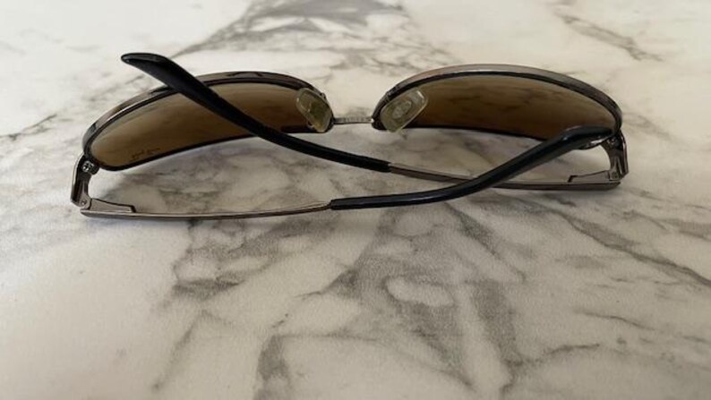 May include: Sunglasses with brown tinted lenses and black arms. The sunglasses have a silver frame and nose pads. The arms are curved and extend from the lenses. The sunglasses are resting on a marble surface.