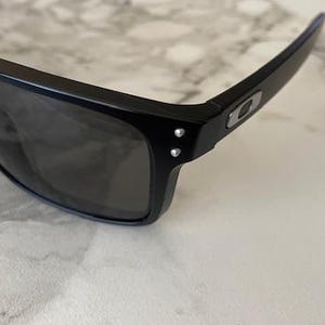 May include: Black sunglasses with dark lenses. The sunglasses have a rectangular frame and a sleek, modern design. The temples feature a silver logo and two small silver rivets. The sunglasses are resting on a white and gray marble surface.