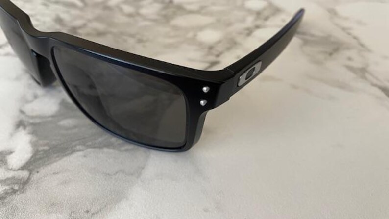 May include: Black sunglasses with dark lenses. The sunglasses have a rectangular frame and a sleek, modern design. The temples feature a silver logo and two small silver rivets. The sunglasses are resting on a white and gray marble surface.