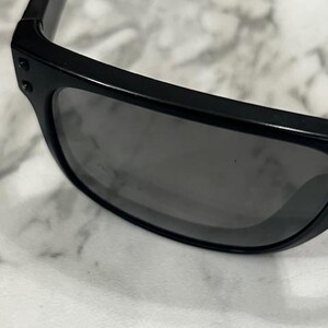 May include: Close-up of black sunglasses with dark gray lenses. The sunglasses have a rectangular frame and are set against a light-colored, textured background. The temples are thick and feature small, round rivets.