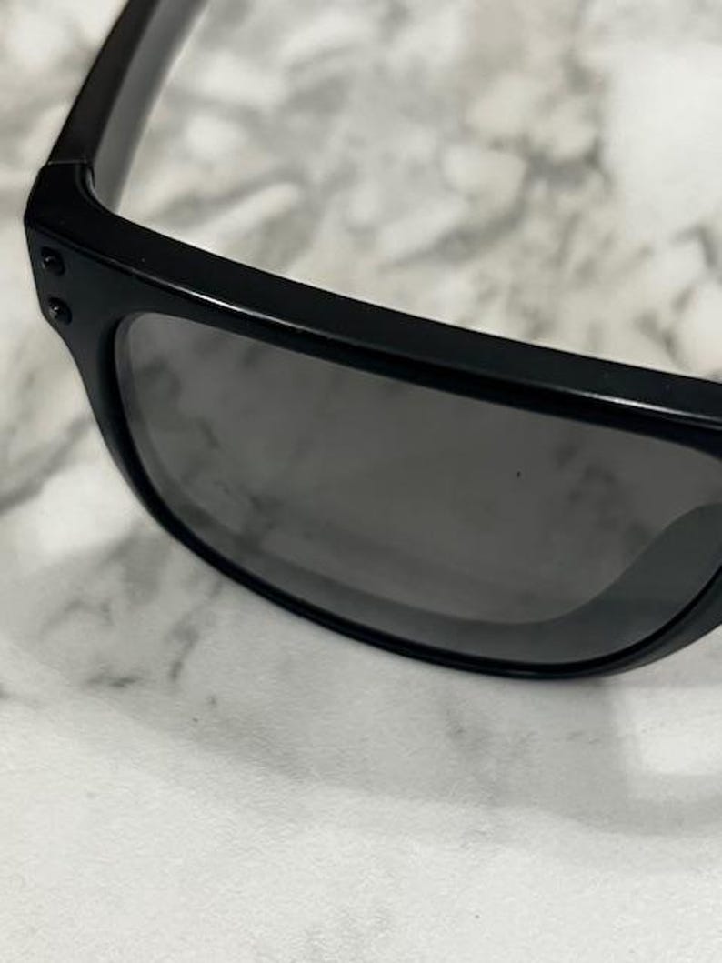 May include: Close-up of black sunglasses with dark gray lenses. The sunglasses have a rectangular frame and are set against a light-colored, textured background. The temples are thick and feature small, round rivets.