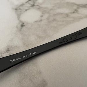 K&ouml;nnte beinhalten: Schwarzer Sonnenbrillenb&uuml;gel mit der Modellnummer OO9238-04, Glasbreite 5,4 cm, Stegbreite 2 cm und B&uuml;gell&auml;nge 13,3 cm. Der B&uuml;gel hat ein dezentes Design und befindet sich vor einem hellen, strukturierten Hintergrund.