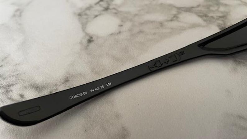 K&ouml;nnte beinhalten: Schwarzer Sonnenbrillenb&uuml;gel mit der Modellnummer OO9238-04, Glasbreite 5,4 cm, Stegbreite 2 cm und B&uuml;gell&auml;nge 13,3 cm. Der B&uuml;gel hat ein dezentes Design und befindet sich vor einem hellen, strukturierten Hintergrund.