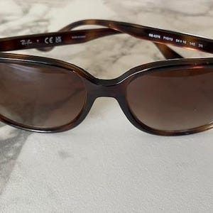 May include: Brown tortoiseshell sunglasses with a cat-eye shape. The Ray-Ban logo is visible on the left lens. The sunglasses have brown-tinted lenses and a dark brown frame. The arms of the sunglasses are also brown.