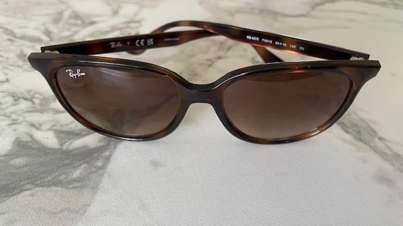 May include: Brown tortoiseshell sunglasses with a cat-eye shape. The Ray-Ban logo is visible on the left lens. The sunglasses have brown-tinted lenses and a dark brown frame. The arms of the sunglasses are also brown.