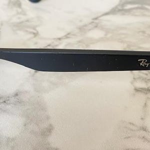 May include: Close-up of black sunglasses with the Ray-Ban logo on the arm. The sunglasses are set against a marble-like surface. The frame is matte black, and the lenses are dark.