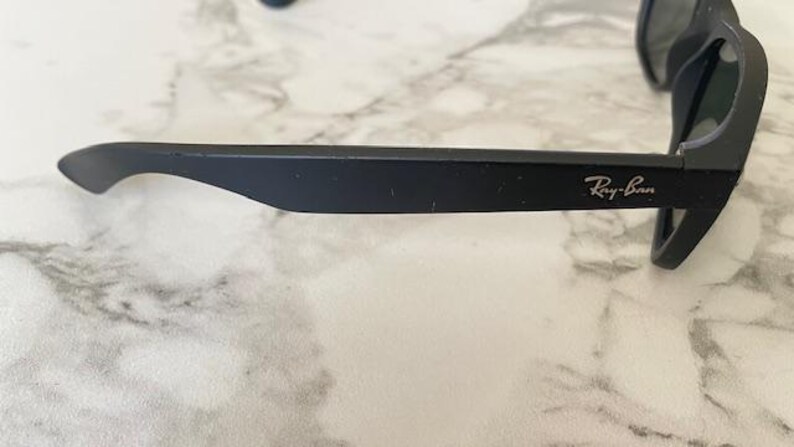 May include: Close-up of black sunglasses with the Ray-Ban logo on the arm. The sunglasses are set against a marble-like surface. The frame is matte black, and the lenses are dark.