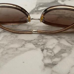 May include: Brown sunglasses with a gold-colored frame and brown lenses. The sunglasses have a rounded shape and are resting on a marble surface. The arms of the sunglasses are a light brown color.