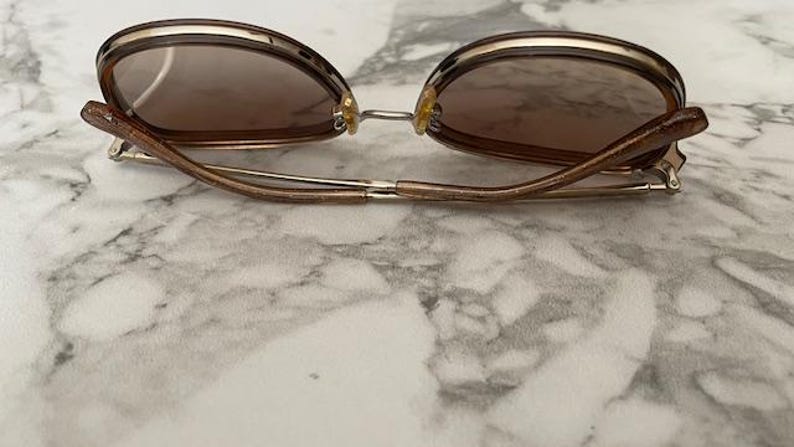 May include: Brown sunglasses with a gold-colored frame and brown lenses. The sunglasses have a rounded shape and are resting on a marble surface. The arms of the sunglasses are a light brown color.