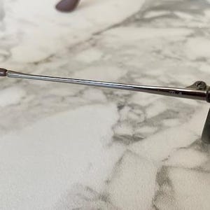 K&ouml;nnte beinhalten: Nahaufnahme einer Sonnenbrille mit braunem Gestell und B&uuml;geln sowie einem silberfarbenen Metallarm. Die Gl&auml;ser sind dunkel gef&auml;rbt. Die Sonnenbrille liegt auf einer Marmoroberfl&auml;che.